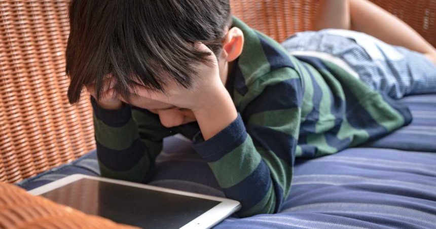 Boy lays on couch with head in hands while using iPad